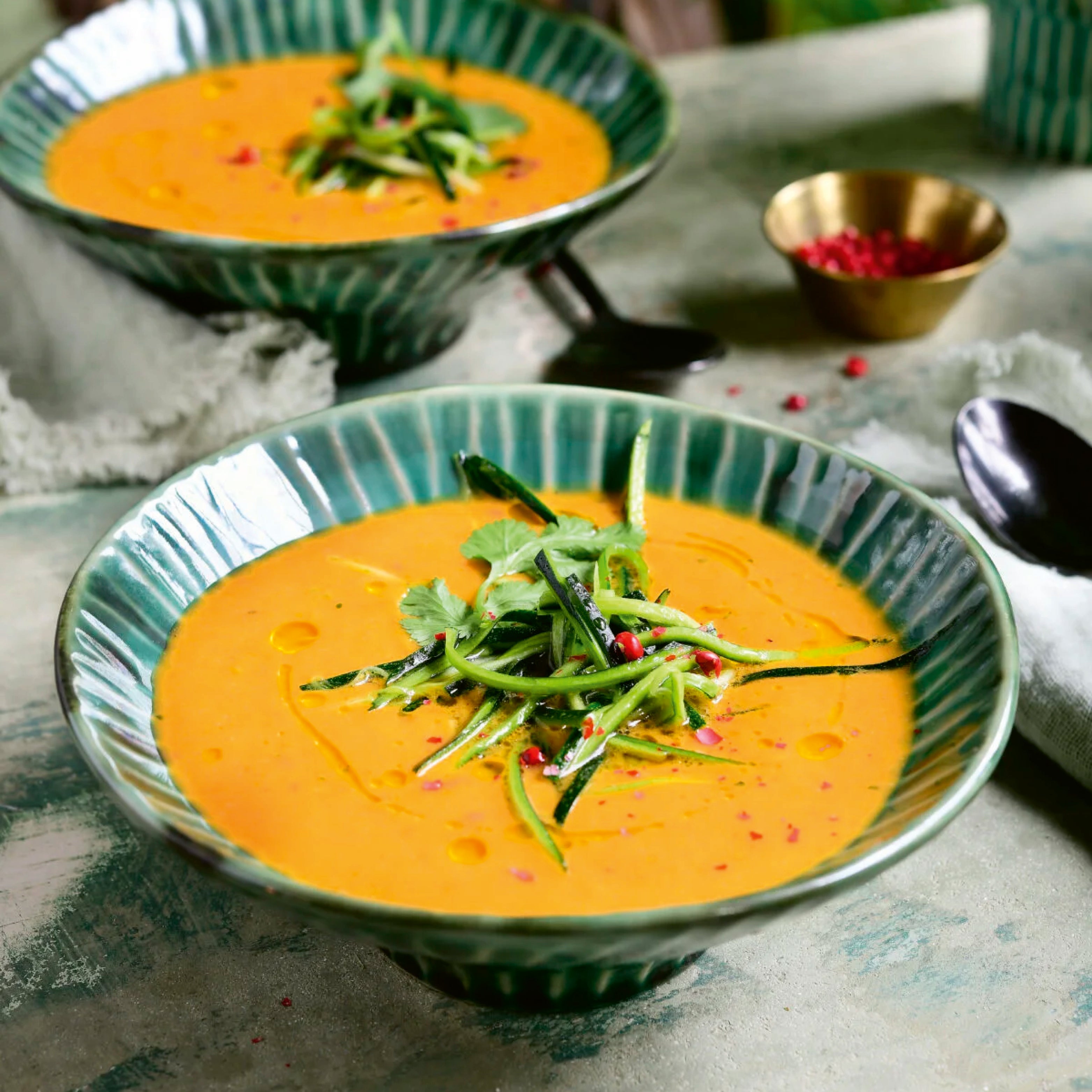 Zwei tiefe, grün glasierte Schalen mit cremiger orangefarbener Suppe, garniert mit feinen Gemüsestreifen und Kräutern, umgeben von Löffeln, Servietten und kleiner Schale – stilvolle Esstisch Dekoration im Gourmet-Look.