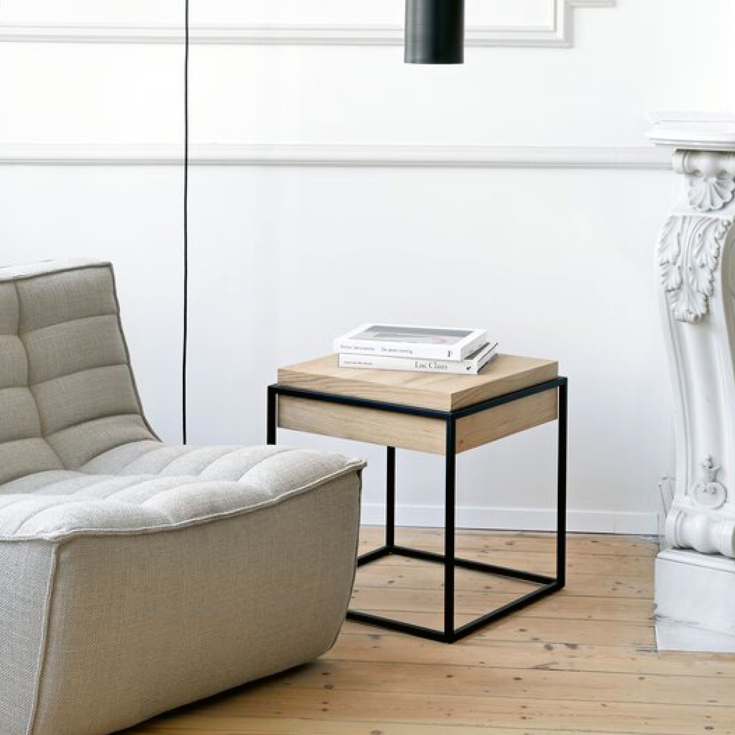 Moderner Beistelltisch im Wohnzimmer: massives Holz mit schwarzem Metallgestell im Kubus-Design, neben einem gepolsterten Loungesessel auf hellem Holzboden – minimalistisches, helles Interieur.