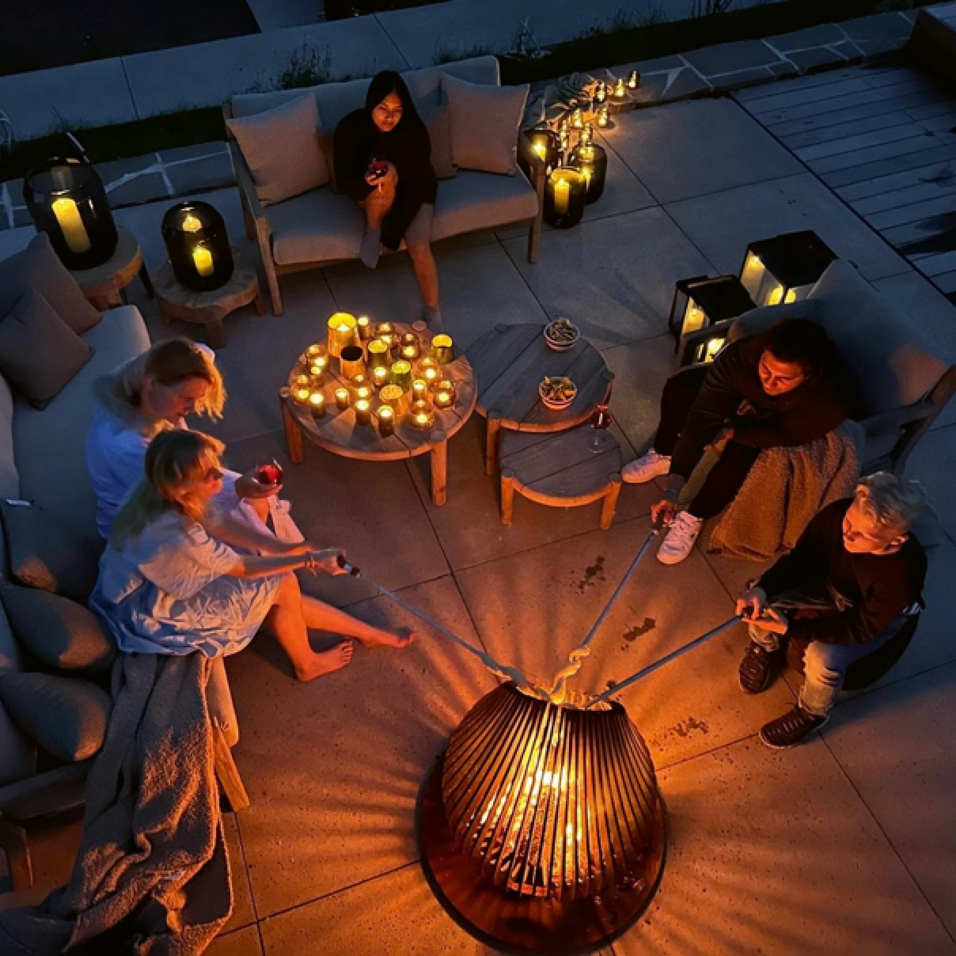 Familie versammelt sich abends um eine Feuerstelle auf der Terrasse, umgeben von Kerzen, Lichtern und gemütlichen Sitzmöbeln – stimmungsvolle Outdoor-Momente mit Kindern.