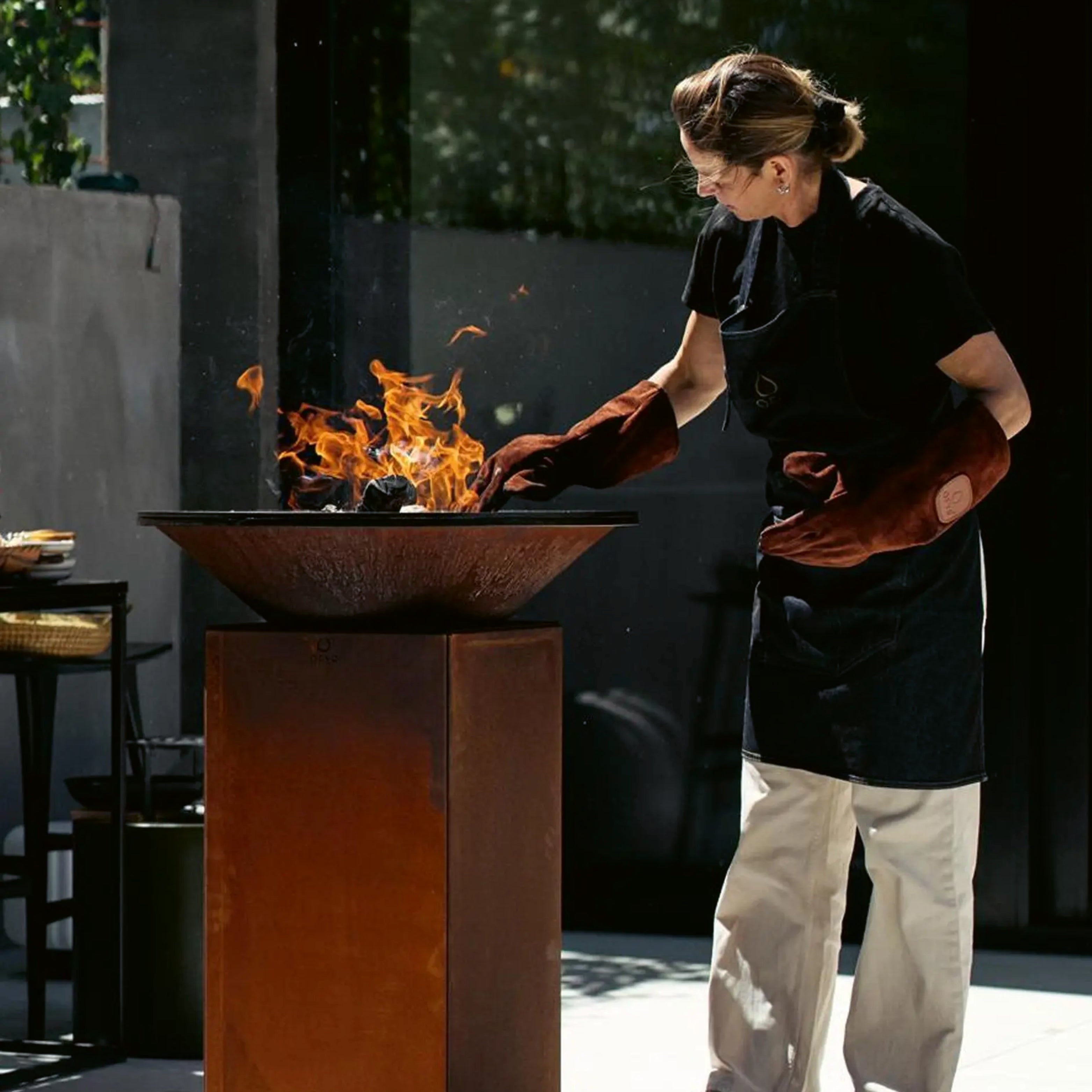 Person grillt an einem rustikalen Designer-Feuergrill mit Cortenstahl-Sockel – modernes Outdoor-Cooking mit Stil und Präzision.