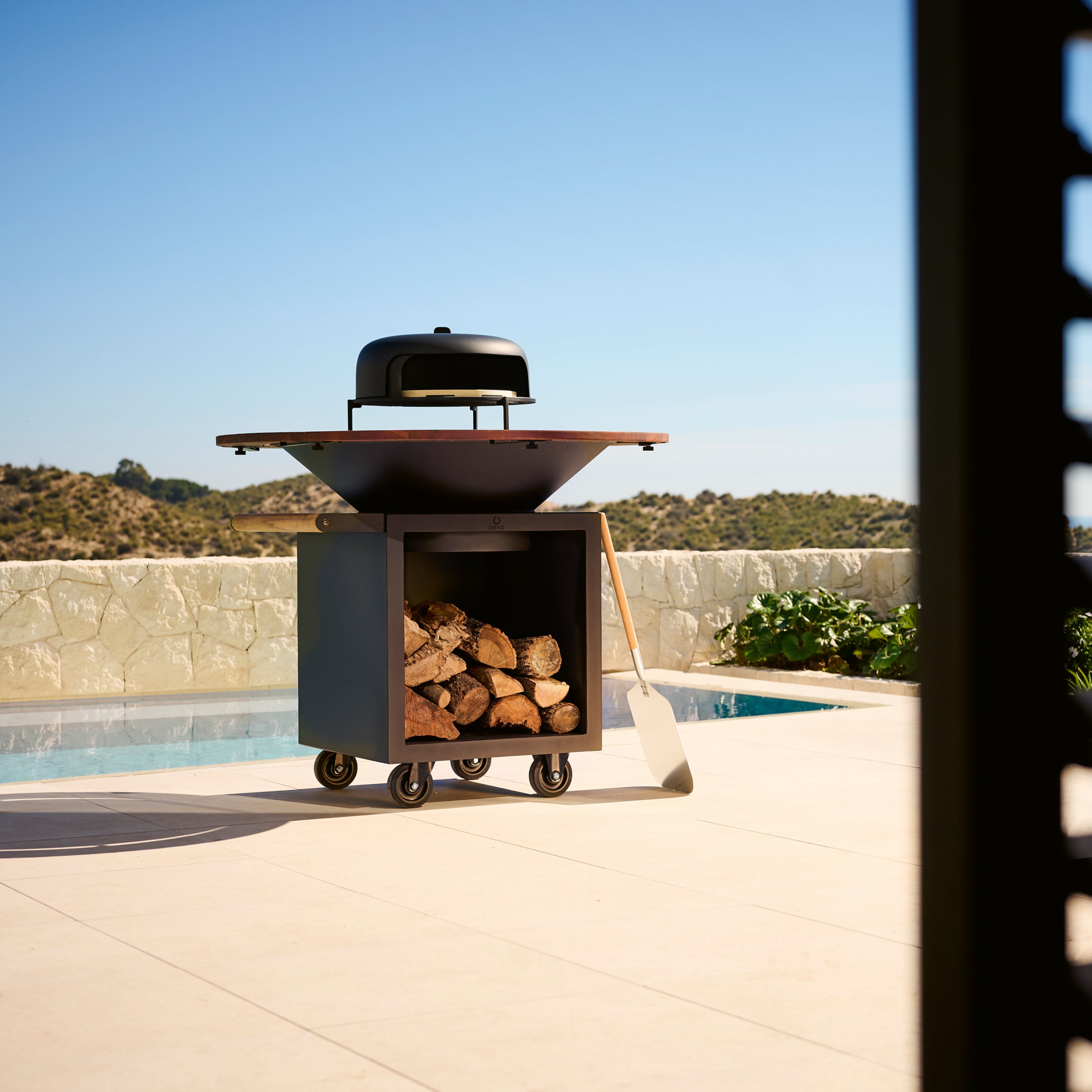 Ofyr Grill mit Holzlager und Pizzaofen auf einer Terrasse, im Hintergrund ein Pool und strahlend hellblauer Himmel – sommerliche Gourmet Homestorys Outdoor-Küche.